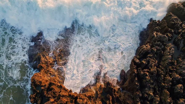 Aerial view stormy ocean waves breaking at volcanic rocks. White spray crashing by huge stones in shallows zoom out. Closeup foamy sea surfs washing dark mountain. Water stream landscape slow motion
