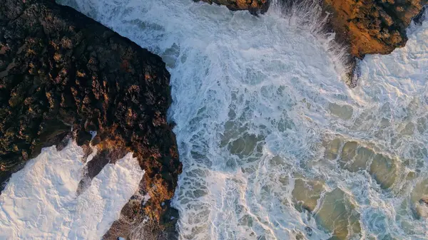 Drone view stormy ocean waves breaking at volcanic coastal. Closeup dramatic foamy sea surfs crashing on stony seashore splashes white froth. Rough clear deep water washing scenic coast slow motion