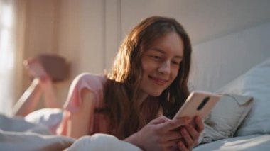 Yatakta sosyal medyada gezinen güzel bir kız. Mutlu genç bir kadın sabahları evinde cep telefonuyla yazı yazıyor. Akıllı telefon mesajları kullanan kaygısız neşeli kadın. Neşeli model görünümlü ekran.