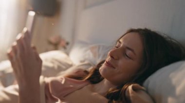 Mutlu kadın daktilo mesajları sabah yakın planda. Neşeli, uykulu kadın sohbeti. Gün ışığında cep telefonuna bak. Rahat güzel kız hücreyi kullanarak rahat bir yatakta uzanıyor. Sakin model, hafta sonunun tadını çıkar.