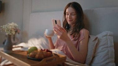 Sabah kadını yatakta bir yudum kahve yudumluyor. Gülümseyen sakin kız internette cep telefonuna bakıyor. Carefree uygulaması müşteri kaydırma hücresi. Gevşemiş güzel kadın hafta sonları kahvaltı yapıyor..