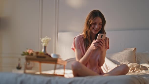 一个快乐的女人在酒店公寓里用智能手机发短信梦幻般的女孩喜欢早上在舒适的床上用早餐桌打手机 美丽而轻松的女性在阳光下浏览社交媒体 — 图库视频影像