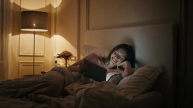 Yatakta tablet izleyen neşeli bir kadın. Akşam yatmadan önce mutlu kadın kahkaha filmi. Uykucu kadın dijital bilgisayara bakıyor ve geceleri komik şovların tadını çıkarıyor. Ev eğlencesi eğlence konsepti