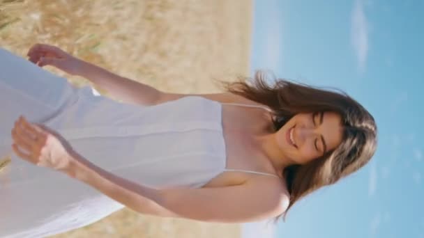 日光の小麦畑を散策するハッピーレディ 垂直に新鮮な笑顔の女性は 自然景観の夏の穀物作物を歩いています カントリーファームランドでカメラに指を向けるホワイトドレスの楽しいビームモデル — ストック動画