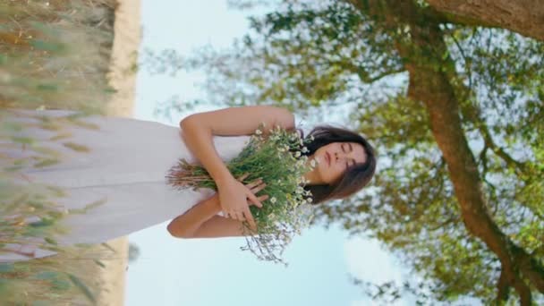 庭の垂直にカモミールの花とポーズするリラックスした女の子 魅力的なロマンチックな女性は風の強い朝の小麦畑に立っているブーケを保持します 田舎の自然の中でリラックスしたブルネット カントリーリビングコンセプト — ストック動画