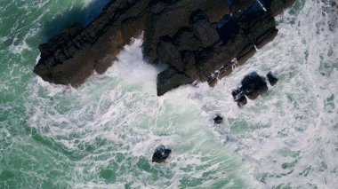 Karanlık taş kayalıklarda köpüren deniz suyu süper yavaş çekimde kırılıyor. Hava manzaralı fırtınalı okyanus, kıyı kayalarını parçalayarak beyaz köpük yapıyor. İnanılmaz doğa tehlikeli kayalık kıyılar güneşli bir gün.