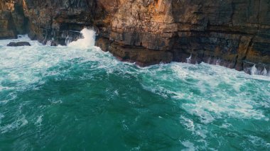 Köpüklü dalgalar kayalık kıyı şeridini ağır çekimde yıkıyor. Fırtınalı derin okyanus sularının üzerindeki tehlikeli kara kıyı uçurumları. Dalgalı deniz kıyısı dağına çarpıyor. Doğa konsepti.