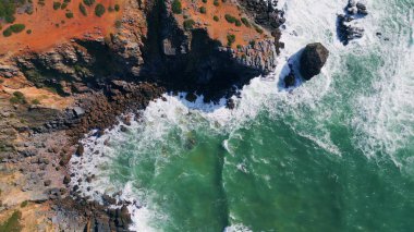 Fırtınalı köpüklü okyanusun üzerinde sinematik manzaralı kayalık bir sahil. İnsansız hava aracı, güçlü deniz dalgalarının tehlikeli volkanik kayalıkları yavaş çekimde vurduğu güneşli deniz manzarasını görüyor. Güzel dalgalar taşlara sıçrıyor.