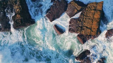 Deniz kıyısındaki kayalıklarda çok güzel fırtınalı deniz köpüğü sıçrıyor. Karanlık kıyı kayalıklarına vuran köpüklü okyanus dalgaları. Güneşli bir gün. Havadan bakıldığında dalgalı deniz taşları suyla yıkanır..