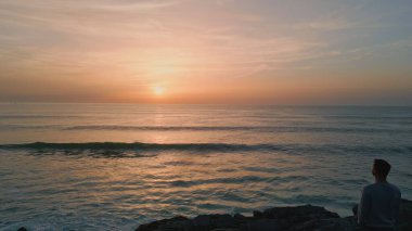 Karanlık uçurumların ardında gün batımını izleyen bir adam silueti. Panoramik şafak deniz manzarası drone görüntüsü. Sonsuz manzaralı okyanus ufku. Pastel pembe ufuk çizgisi. Portakal rengi günbatımı, sakin suyla yansıyor.