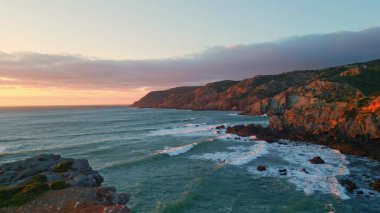Volkanik kayalıklar, günbatımı okyanus manzarasıyla buluşuyor. Güzel köpüklü deniz dalgaları sert kayalık sahil yamaçlarına süper yavaş çekimde yuvarlanıyor. Akşam deniz suyu kıyıları köpükle kaplıyor.