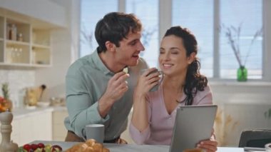 Kahkaha atan çiftler kahvaltıda dijital tabletleri yakın çekim olan rahat mutfağın keyfini çıkarıyorlar. Neşeli genç eşler sabah birlikte eğleniyorlar. Neşeli kadın elinde bilgisayarla kocasıyla kahve içiyor.