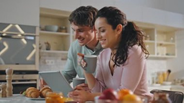 Romantik çift aile kahvaltısı için internetten alışveriş yapıyor. Mutlu gülen eşler kruvasanla kahve içerken modern mutfakta tablet bilgisayar ekranına bakarlar. Dijital aygıt kullanan neşeli çift
