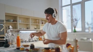 Modern mutfakta kablosuz kulaklıkla müzik dinleyen bir adam. Kaygısız gülümseyen adam kahvaltıyı kulaklıkla hazırlıyor dans ediyor ve şarkı söylüyor. Mutlu yakışıklı, tatlı tost yaparken eğleniyor.