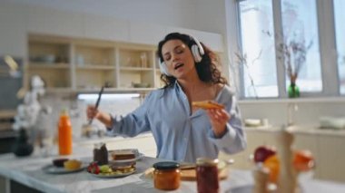 Kulaklıklı kız modern mutfakta kahvaltı hazırlıyor. Kaygısız mutlu kadın kulaklıkla en sevdiği şarkıyı söyleyerek evde çikolata tostu hazırlıyor. Kaygısız esmer dans müziği seviyor..