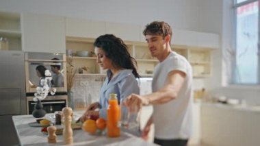 Romantik çiftler rahat ev yemeklerinde yemek yapmayı severler. Güzel mutlu kız arkadaş mutfak masasında yemek hazırlıyor. Gülümseyen erkek arkadaş portakal suyu için gözlük alıyor. Sevgili eşlerin aile hafta sonunu geçirmesi.