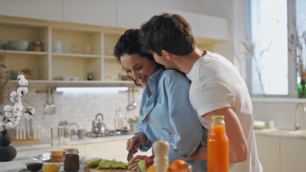 幸福的已婚夫妇在家里做饭 快乐温柔的丈夫拥抱美丽的妻子 切新鲜的水果作为健康的早餐 可爱的配偶们在舒适的厨房里开心地笑着 — 图库视频影像