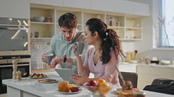 丈夫与妻子吻别继续工作 快乐微笑的夫妻在工作前一起在现代化厨房喝咖啡 松驰的漂亮女人手握平板电脑坐在台面 — 图库视频影像