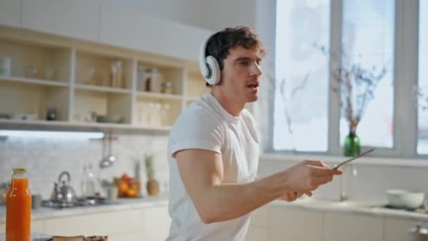 一个戴着无线耳机在厨房里听音乐的家伙走过来了 快乐悠闲的男人在耳机里跳舞 准备美味的烤面包 准备在舒适的烹饪室内吃早餐 Melomaniac用耳机烹调 — 图库视频影像