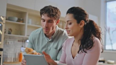 Mutlu eşler sıcacık mutfakta tabletle kahvaltı yapıyor. Rahatlamış, gülümseyen ve kahveli kruvasan yiyen çift evde birlikte bilgisayar ekranına bakıyorlar. Genç aile çevrimiçi alışveriş yapıyor