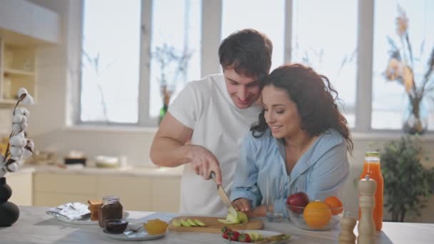 可爱的配偶们正在为健康的早餐准备近餐 快乐的年轻人在厨房台面切新鲜苹果 美丽而快乐的女人看着用爱心烹调丈夫 — 图库视频影像