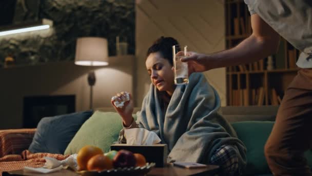 丈夫照顾生病的妻子 躺在温暖的格子布沙发上 有爱心的男人把水杯带给生病的女人 支持在家里拥抱 流鼻涕 打喷嚏 患感冒的小女孩 — 图库视频影像