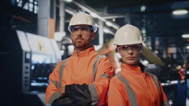均一なクローズアップで金属製の生産に立っているカップルの工場技術者 自信を持って製造施設でポーズする2人のエンジニア ヘルメットにおける深刻な産業労働者の肖像 — ストック動画