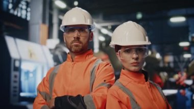 Fabrika mühendisleri koruyucu kask üniformalı imalat atölyesinde poz veriyorlar. Endüstriyel makinelerin yanında duran ciddi fabrika işletmecileri. Kendine güvenen iki teknisyen kameraya bakıyor..