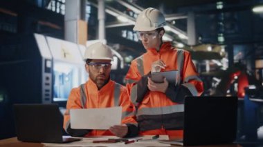 İki fabrika çalışanı atölyedeki endüstriyel üretim belgelerini kontrol ediyor. Erkek mühendis, tabletli kadın teknisyene teknik evrakları gösteriyor. Üniformalı birkaç fabrika işçisi işbirliği yapıyor..