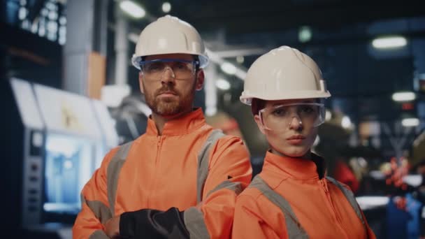 保護ヘルメットのユニフォームの製造工場でポーズするプラントエンジニアが閉じます 産業機械の近くに立っている真剣なカップルの工場オペレータ カメラを見ている2人の技術者が自信を持って — ストック動画