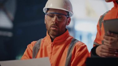 Fabrika işçileri üretim tesislerinin teknik belgelerini inceliyor. Profesyonel bir zanaatkar tabletli bir kadına düzen formu gösteriyor. Üniformalı iki teknisyen üretim bilgilerini tartışıyor 