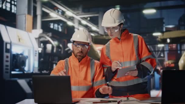 チームの工場労働者は 工業製品のラップトップコンピュータを見ながら話しています ヘルメットユニフォームの2人のエンジニアがイノベーション生産について話し合います カップル専門の専門家が植物に取り組む — ストック動画
