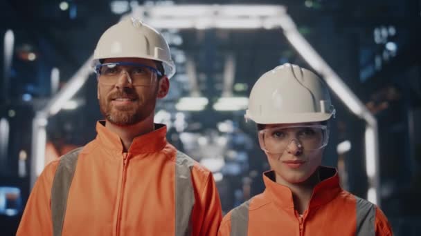 保護ヘルメットを着用する近代的な施設でポーズするカップル成功した工場エンジニア 金属工房に立っている2人の幸せな技術者 カメラに均一笑顔のプルードプラント労働者 — ストック動画
