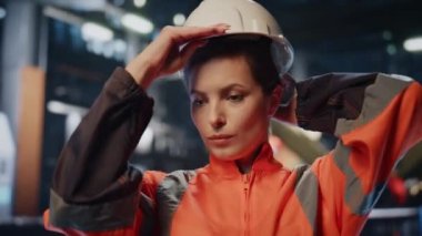 Ağır endüstri çalışanı kameraya gülümseyen koruyucu bir kask takıyor. Ön üretim makinelerinde poz veren profesyonel endüstriyel mühendis kadın. Kendine güvenen kadın zanaatkar ayakta duruyor.