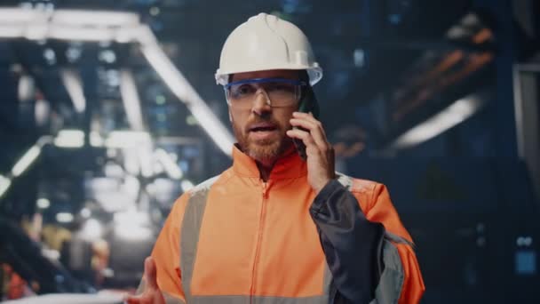 金属工場の作業問題を解決するスマートフォンを話す神経質な産業労働者 携帯電話で問題を議論する真剣なエンジニア ヘルメットの制服電話のひげ付き技術者 — ストック動画