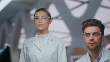 Modern klinikte kameraya bakan mutlu kimyagerlerin portresi. Gelişmiş laboratuarda poz veren gülümseyen genç bilim adamları takım çalışmasının keyfini çıkarıyorlar. Tıp üniformalı iki profesyonel araştırmacı birlikte çalışırlar.