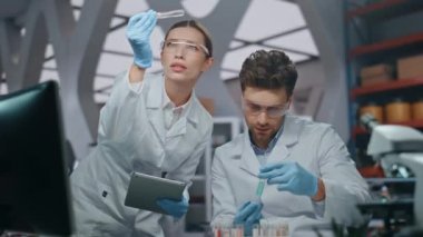Biyoteknoloji laboratuvarında araştırma yapan profesyonel bilimadamları yakın çekim yapıyorlar. Gözlüklü iki araştırmacı birlikte test tüplerine bakıyor. Elinde tablet olan kadın, erkek meslektaşıyla aşı üzerinde çalışıyor.