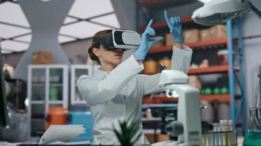 Modern laboratuvarda sanal gerçeklik teknolojisiyle çalışan gözlüklü bilim adamı. VR gözlüklü kadın doktor arayüze elle dokunuyor. Tıbbi araştırmacı, geliştirilmiş gerçekliği analiz için kullanıyor..