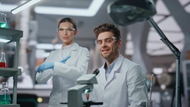 在现代化的实验室里 笑着看着相机的科学家们非常接近了 创新诊所的专业科学研究人员穿着医疗制服摆姿势 高级实验室快乐的两位生物化学家的画像 — 图库视频影像
