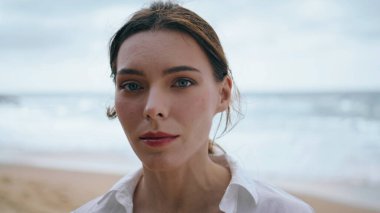 Güzel, nazik kadın yüzü kameraya dikkatlice bakıyor. Deniz dalgalarına yakından bakıyor. Bulutlu sahilde tek başına beyaz gömlek giyen çekici ve düşünceli bir kızın portresi. Deniz kıyısında poz veren sakin bir bayan.