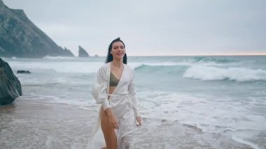 Mutlu kadın turist bulutlu deniz kıyısında gülümseyen kamerayla dinleniyor. Çekici kaygısız kız okyanus dalgalarında beyaz pareo giyerek yürüyor. Güzel genç bayan Rocky sahilinde tatilin tadını çıkarın..
