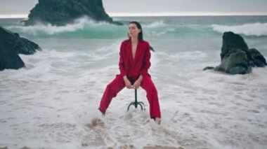 Sahilde oturan zarif güzel bayan köpüklü dalgalarla çıplak bacakları yıkadı. Kaya grisi dramatik okyanus manzarasının yanındaki sandalyede poz veren kendine güvenen çekici bir model. Kırmızı şık takım elbiseli şehvetli bir genç kadın..