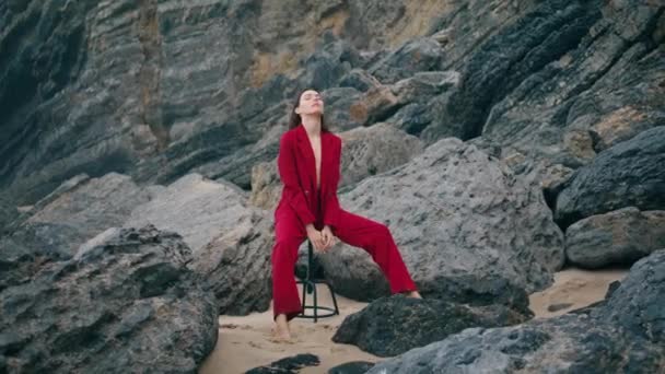 穏やかなリラックスした女性は エレガントな赤いスーツを着てビーチ崖にポーズします ロッキー海岸の椅子に座っている魅力的なモデル スタイリッシュな豪華な女の子は暗い自然にリラックスします スタイルコンセプト — ストック動画