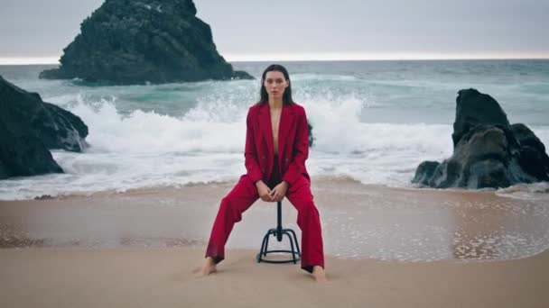赤いスーツを着た暗い海岸にカメラをポーズするエレガントな豪華なモデル 海の波の前で椅子に座っている自信のある真面目な女性 穏やかなリラックスした女の子は海の自然を楽しんでいます スタイルコンセプト — ストック動画