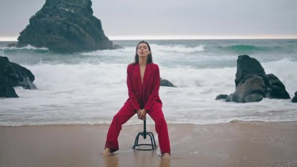 沉着优雅的女孩在汹涌的浪花前坐在椅子上 享受着暗淡的海景 自信而严肃的女人穿着时髦的红色西服在多云的海滩上摆姿势 性感时尚的女士感到宁静 — 图库视频影像
