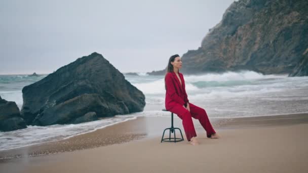 赤いセクシーなスーツの曇った日にビーチに座っている豪華なスタイリッシュな女性 ロッキーな嵐の海岸で椅子にポーズするセンセーショナルな魅力的な少女 暗い海岸で落ち着いた思慮深い孤独に見える優雅な若い女性 — ストック動画