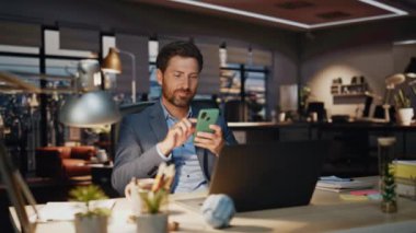 Akşam lambası ofisinde cep telefonu mesajı yazan gülümseyen CEO. Masada akıllı telefon e-postalarını okuyan mutlu bir işadamı. Pozitif adam panoramik pencerelerde cep telefonu kullanırken mutlu hissediyor.
