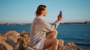 Sahil kızı gün ışığında akıllı telefonuyla fotoğraf çekiyor. Gün batımında cep telefonuyla nehir kıyısında dinlenen güzel bir kız. Sakin bir manken, sosyal medya için deniz fotoğrafı yapar. Dijital yaşam tarzı kavramı