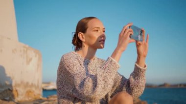 Shore Sunset 'te yakın çekim yapan deniz modeli videosu. Güzel bir kız göl kenarında dinleniyor ve akşam güneşinde cep telefonuyla fotoğraf çekiyor. Yaz tatilinde akıllı telefon kullanan kaygısız genç kadın.
