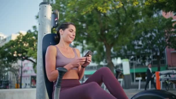 有吸引力的运动员在手机旋转踏板上回答信息的自行车机械设备 积极的女运动员一个人在城市露天体育馆检查手机训练 健康生活方式概念 — 图库视频影像